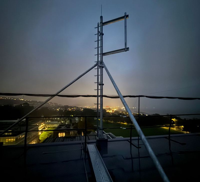 Tripod antenna pole with cable tray on a rooftop at dusk, town lights behind